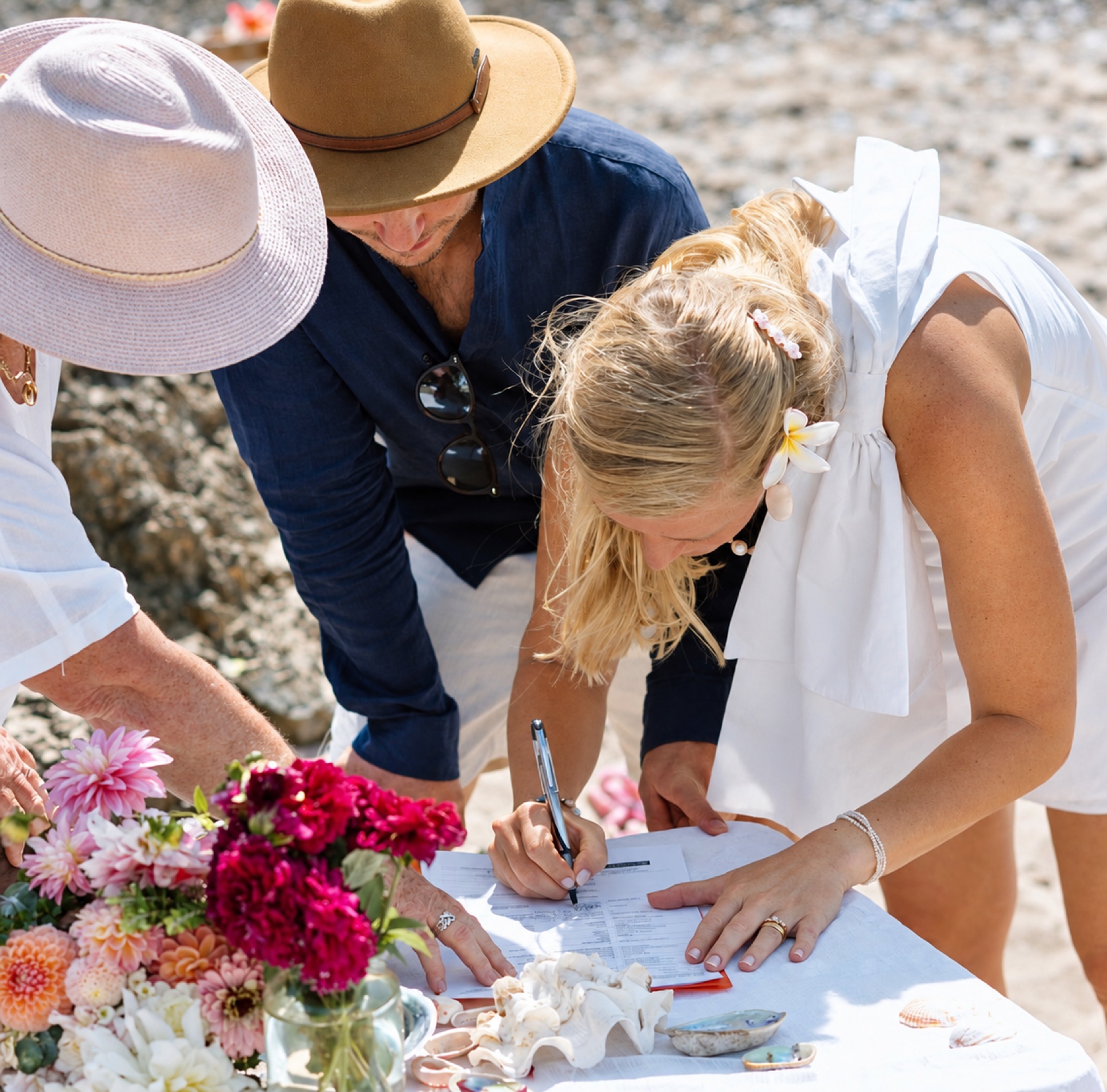 A bride signing the marriage paperwork at a flower-covered table on the beach