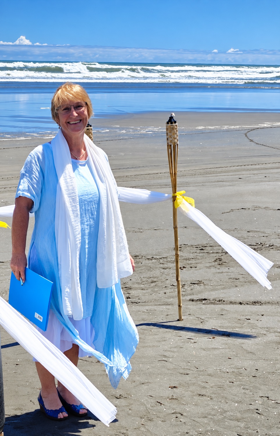 Chris on a black sand beach in a pale blue dress, holding a blue folder beside white and yellow ribbons