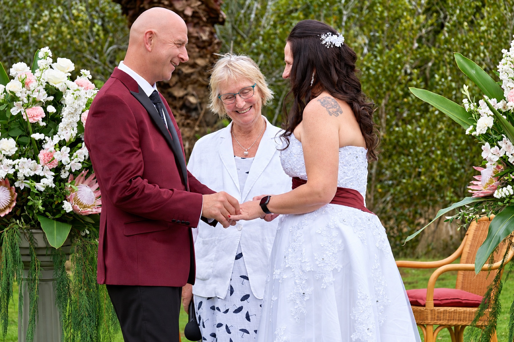 Chris leading a garden ceremony with a groom in a red suit and a bride in white